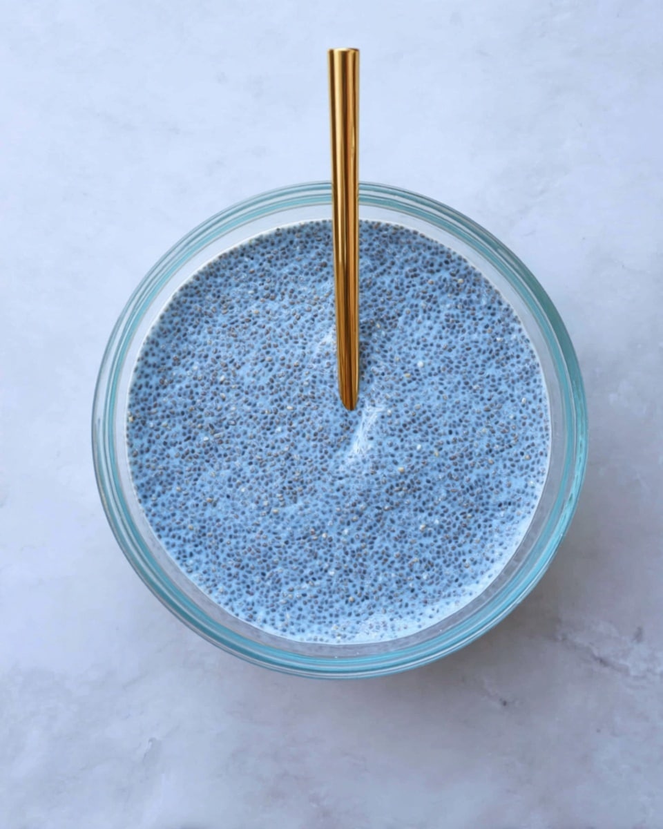 A clear glass bowl filled with a single smooth layer of light blue chia pudding, showing evenly spread tiny chia seeds throughout the mixture. A golden spoon stands upright in the center of the bowl. The bowl is placed on a white marbled surface, with a clean and minimal look. photo taken with an iphone --ar 4:5 --v 7