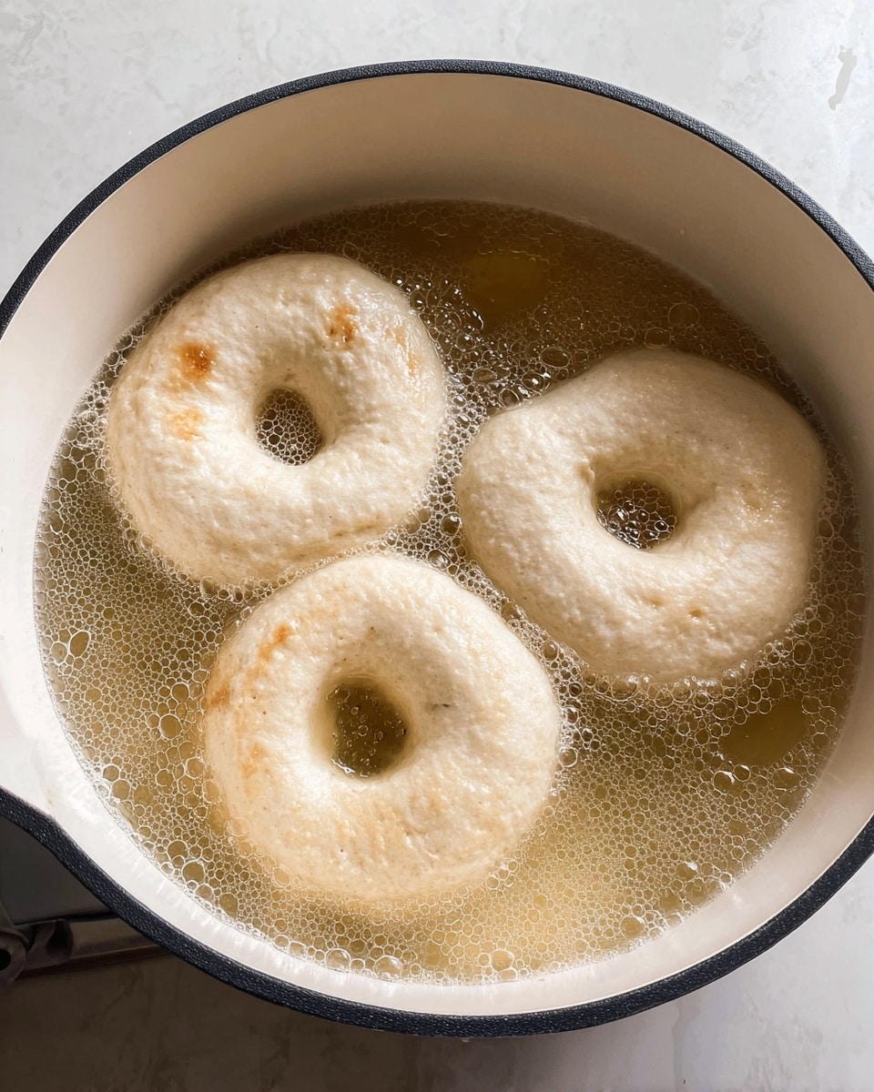 Three pale dough rings float in bubbling light golden oil inside a white pot with a black rim. The dough rings have a soft and slightly uneven surface with a clear hole in the middle of each. Tiny bubbles gather around the edges of the rings and the pot’s inner wall shows light brown marks near the top. The scene is set on a white marbled texture. photo taken with an iphone --ar 4:5 --v 7