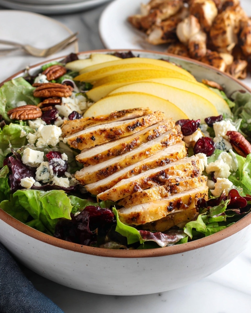 A white bowl filled with layers of fresh green and purple leafy lettuce, topped with layered slices of yellow pear fanned out on one side, scattered pieces of white cheese, deep brown pecans, and small bits of dark red dried cranberries. On top, there is a thick layer of grilled chicken sliced into several pieces showing a golden-brown, slightly peppered surface. The bowl sits on a white marbled surface with a blurred white plate holding chicken pieces in the background. Photo taken with an iphone --ar 4:5 --v 7