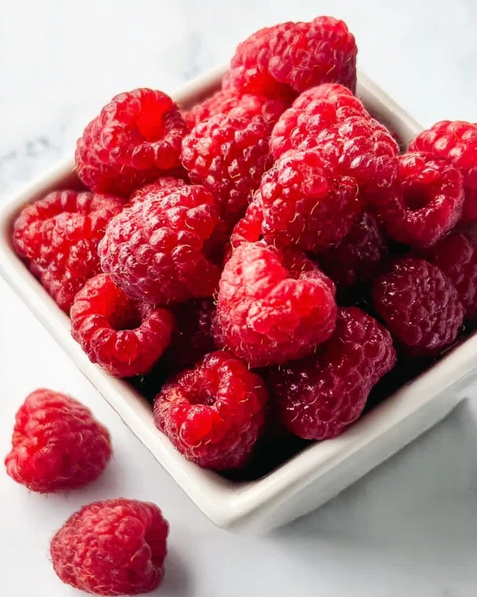 A close-up of a small white square bowl filled with bright red raspberries that have a shiny, bumpy texture. The raspberries are piled high, with some round and some more heart-shaped at the top edges of the bowl. Two raspberries lie next to the bowl on the white marbled surface, adding depth to the image. The lighting highlights the fresh, juicy texture of the fruit, creating small shiny spots on the raspberries. photo taken with an iphone --ar 4:5 --v 7