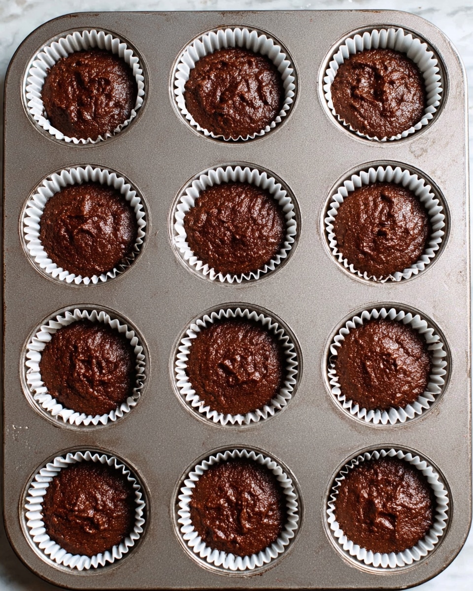 A dark brown chocolate batter is placed inside twelve white paper liners, each sitting evenly in a metal muffin tray. The batter looks thick and slightly rough on the surface with small uneven patches, filling each cup about three-quarters full. The tray shows a bit of shine and smooth texture, resting on a white marbled surface. photo taken with an iphone --ar 4:5 --v 7