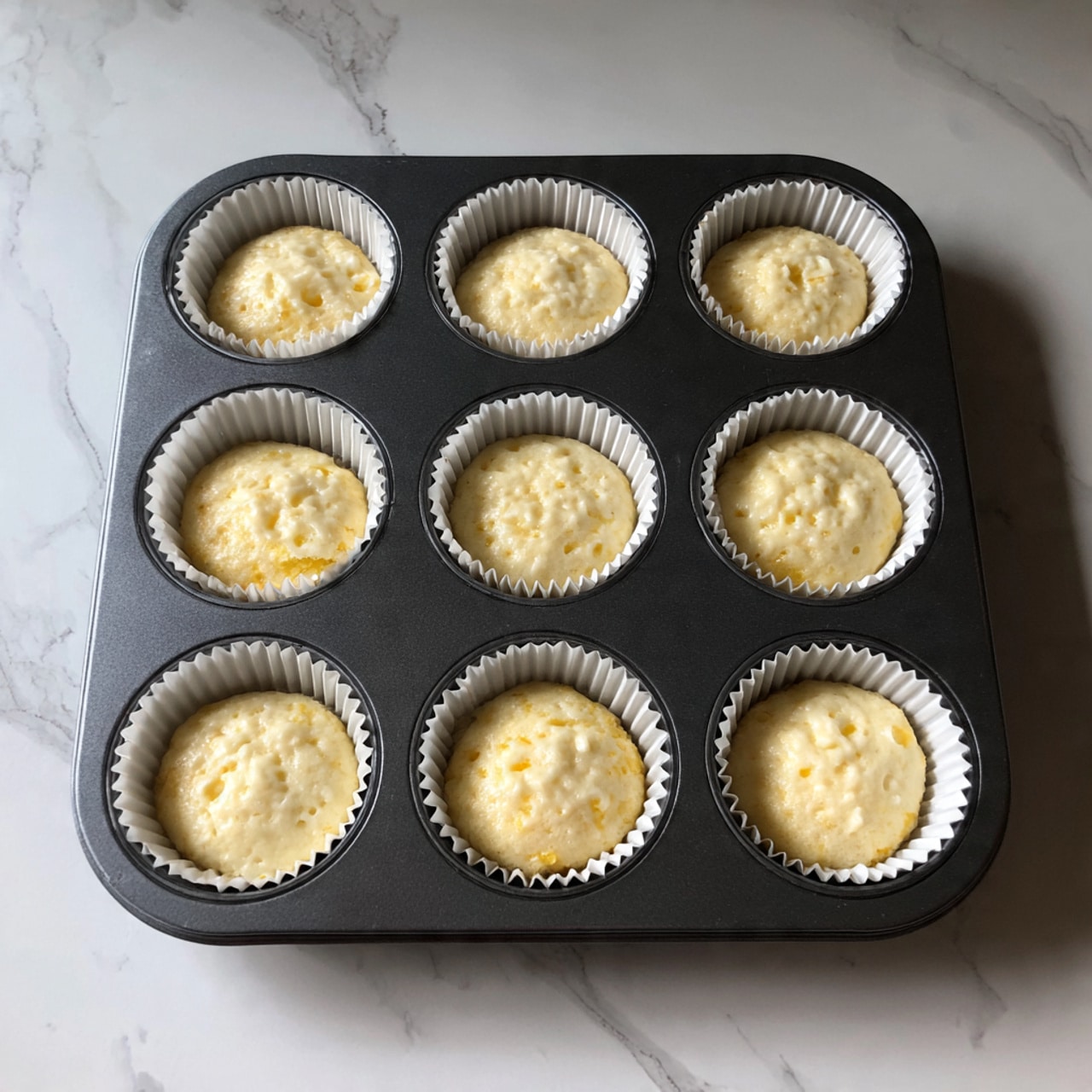 The image shows a close-up view of a dark gray muffin tray filled with nine muffins in white paper liners. Each muffin has a light yellow batter with a smooth, slightly bumpy texture. The muffins sit evenly in the tray's round cups, showing soft tops with small air bubbles visible on the surface. The tray is placed on a white marbled surface, creating a clean and bright background. Photo taken with an iphone --ar 4:5 --v 7