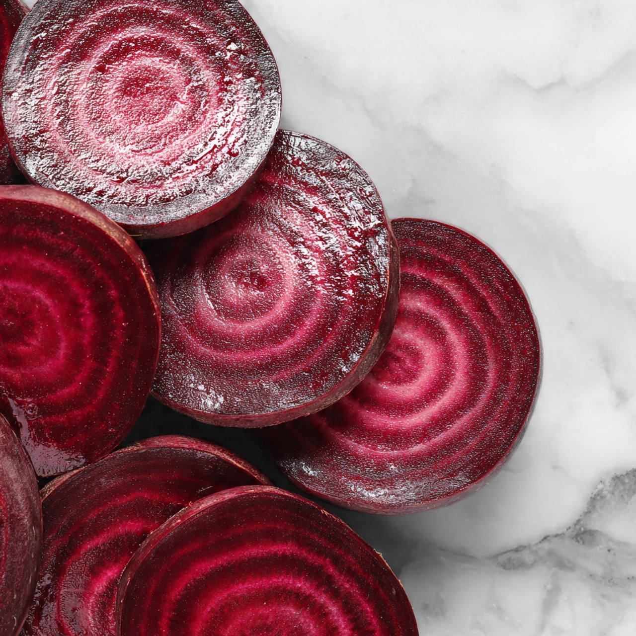 This image shows several rows of thin, round slices of bright red beetroot placed closely together on a dark surface. The slices reveal a smooth, moist texture with concentric circle patterns in varying shades of deep red and burgundy, giving a rich and fresh look. The slices slightly overlap each other, creating a layered effect with a shiny finish from the moisture. The background is replaced with a white marbled texture to highlight the vibrant colors of the beetroot. Photo taken with an iphone --ar 4:5 --v 7