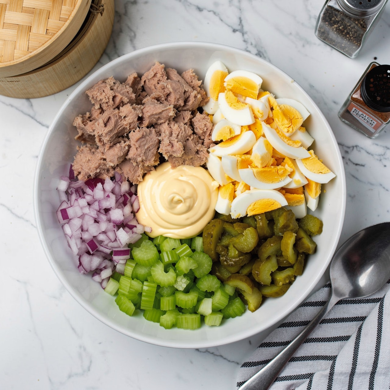 A white bowl sits on a white marbled surface, filled with five separate layers of ingredients. At the top left, there is a thick layer of light brown tuna. At the top right, a pile of chopped boiled eggs with bright yellow-orange yolks contrasts with the white of the egg whites. Below the eggs, there is a layer of chopped green pickles. At the bottom center, there is a bright green layer of chopped celery. Finally, at the bottom left, there is a small pile of finely diced red onions. In the middle of the bowl, a dollop of creamy light yellow mayonnaise sits slightly overlapping the tuna and chopped eggs. Near the bowl, there is a wooden container with a bamboo lid that is slightly open and a small clear glass bottle labeled