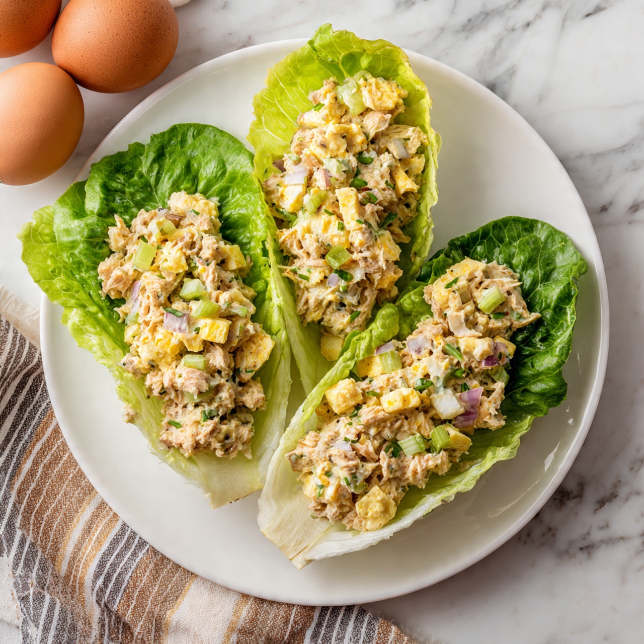 The dish shows three bright green romaine lettuce leaves laid side by side on a white plate, each leaf holding a scoop of chunky salad made of chopped hard-boiled eggs, light orange tuna or chicken, small pieces of celery, and bits of red onion, giving a mix of creamy textures and light colors like yellow, pale green, and red. The plate is set on a white marbled surface with two brown eggs and a striped cloth nearby, adding to the kitchen setting. Photo taken with an iphone --ar 4:5 --v 7