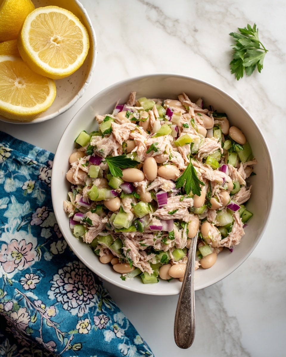 A white bowl is filled with a mixed salad containing three main layers: soft light pink shredded tuna as the base, scattered pale beige beans on top, and small chopped pieces of green celery and purple-red onion throughout. Bright green parsley leaves are sprinkled on the salad, adding fresh color contrast. A silver spoon holds some salad inside the bowl, resting slightly on the right side. To the top left of the frame, a white bowl contains three lemon halves, showing bright yellow peel and juice. A blue patterned cloth with light floral designs lies beside the bowl on the white marbled surface, adding texture. Photo taken with an iphone --ar 4:5 --v 7