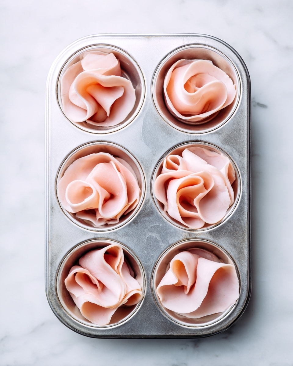 A silver muffin tray with four round cups is shown lined with thin slices of pink ham, each slice draped loosely in a bowl shape with soft curls and folds creating a layered look inside the cups. The tray rests on a white marbled surface that adds a clean, bright background contrast to the pale pink ham and shiny metal of the tray. photo taken with an iphone --ar 4:5 --v 7