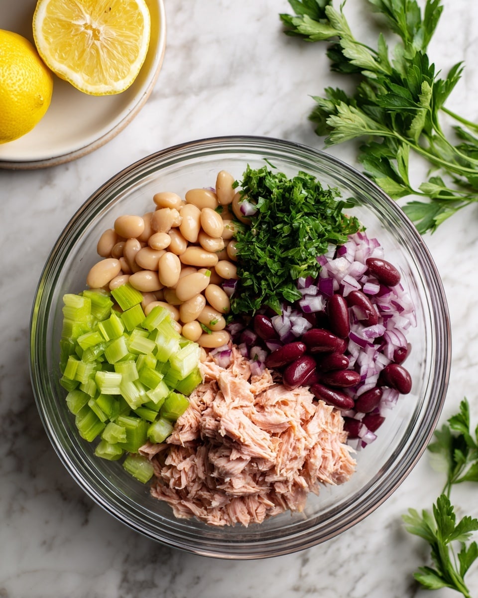 A clear glass bowl holds five layers of ingredients arranged in sections: in the bottom right, light pink shredded tuna with a soft and flaky texture; above it, bright green chopped celery cubes with a crisp look; to the left of celery, pale orange-beige cannellini beans with a smooth, round surface; next to the beans, finely chopped purple-red onions with visible white pieces; and on the top right, fresh dark green flat-leaf parsley sprigs with a leafy texture. Behind the bowl to the upper left is a white plate with two yellow lemon halves. The bowl is placed on a white marbled surface. Photo taken with an iphone --ar 4:5 --v 7