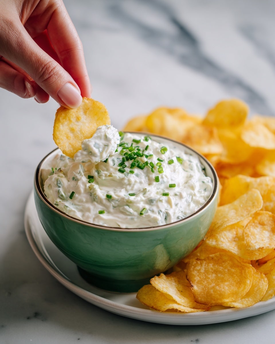 A green bowl filled with creamy white dip mixed with small green chive pieces, topped with more chopped chives in the middle, is placed on a round white plate next to a pile of yellow potato chips. A woman's hand is dipping one chip into the thick, textured dip. The scene is set on a white marbled surface. photo taken with an iphone --ar 4:5 --v 7