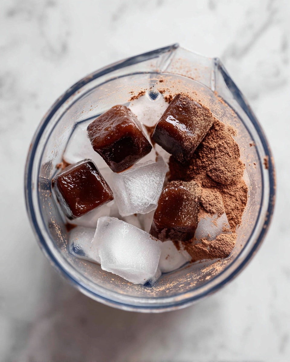 Inside a clear blender, there are several layers: at the bottom, white ice cubes with clear edges, sitting beneath three large dark brown coffee ice cubes with a smooth glossy surface, and a layer of fine light brown powder covering part of the coffee cubes and ice, creating a mix of textures. The blender is on a white marbled surface with slight coffee bean bits around it. Photo taken with an iphone --ar 4:5 --v 7