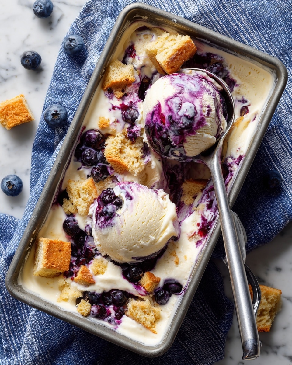 A rectangular gray metal container holds creamy white ice cream with swirls of deep purple blueberry sauce mixed throughout. Scattered on top are several pieces of light golden flaky crust, adding texture and color contrast. A silver ice cream scoop lifts a rounded portion, showing the mix of white ice cream, purple swirls, and crust chunks. The container sits on a blue and white striped cloth, placed on a surface with a white marbled texture. Small blueberries and crust pieces are scattered around. Photo taken with an iphone --ar 4:5 --v 7