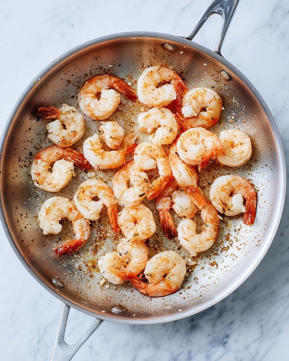 The image shows a round stainless steel frying pan with 19 cooked shrimp inside. The shrimp are arranged randomly, all peeled and curved in shape with a mix of pink and white colors. The pan has some browned bits and small oil spots scattered around, showing it has been used to cook. The background is a white marbled surface. photo taken with an iphone --ar 4:5 --v 7