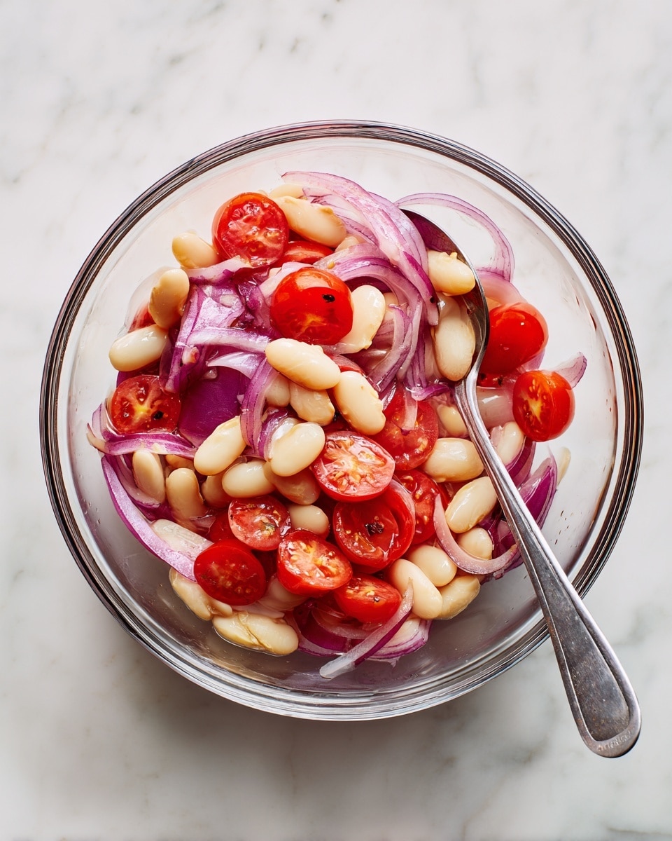 A clear glass bowl holds a simple salad with three main layers: white beans as the base layer, thinly sliced rings of purple-red onion scattered through and on top, and bright red cherry tomato pieces placed evenly around. The textures show the beans as smooth and oval, the onion rings as thin and slightly translucent, and the tomato pieces as juicy with soft edges. A silver spoon is partly inside the bowl, resting near the right edge. The bowl is on a white marbled surface that adds a clean and fresh look. photo taken with an iphone --ar 4:5 --v 7