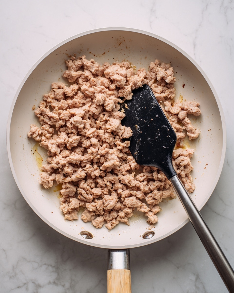 A white pan filled with cooked ground meat pieces that are light brown and crumbly, spread evenly across the pan's surface. A black spatula with a metal handle is placed in the pan, partly under the meat, with visible droplets of oil or moisture on the spatula's surface. The pan handle has a light wood grip, and the background shows a white marbled texture. photo taken with an iphone --ar 4:5 --v 7