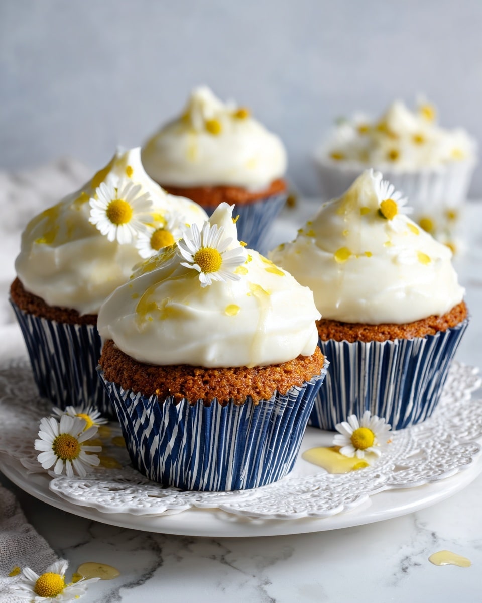 The image shows five cupcakes in blue paper liners on a large white plate with a delicate raised edge. Each cupcake has one thick layer of fluffy white frosting, piled high with soft peaks and crowned with small yellow and white chamomile flowers. The cupcakes have a brownish cake base visible beneath the frosting. Around the plate are scattered chamomile flowers and green stems along with thin drops of honey glistening on the white marbled surface under the plate. The scene looks bright and fresh with a soft focus on the cupcakes and a clean, white marbled background photo taken with an iphone --ar 4:5 --v 7