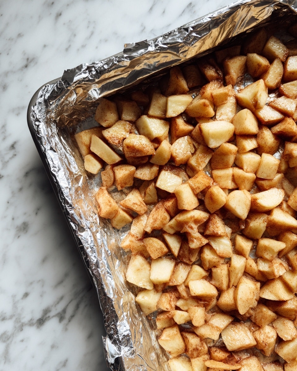 The image shows a close-up of a baking tray lined with aluminum foil, filled with small pieces of light tan and slightly browned apple chunks. The apple pieces are spread evenly across the tray, creating a single thick layer. The background is a white marbled surface, and the overall look is warm and textured, highlighting the cooked apples. Photo taken with an iphone --ar 4:5 --v 7