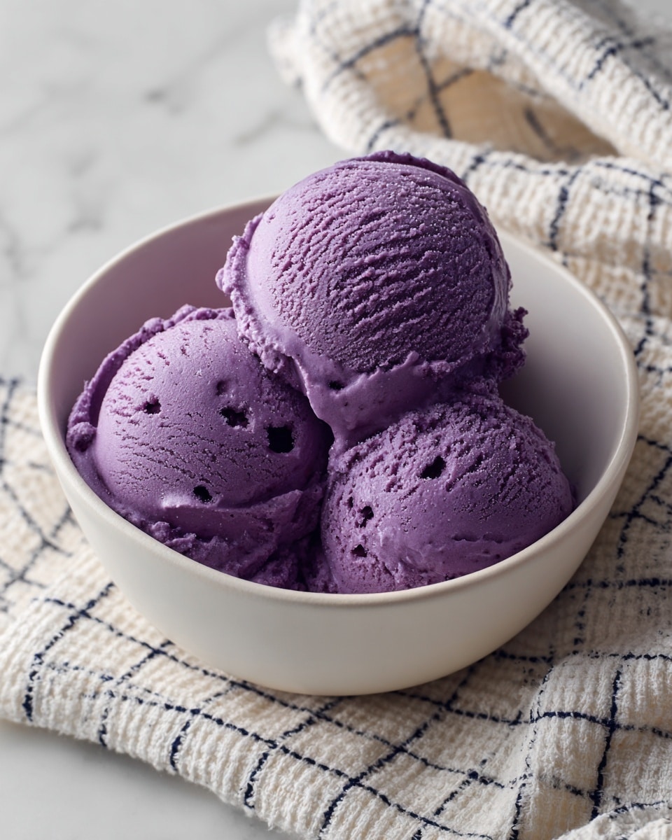 The image shows a white bowl filled with three scoops of smooth, creamy purple ice cream slightly melting at the edges. The ice cream has a rich violet color with a slightly shiny texture and some small specks visible throughout. The bowl sits on a white marbled surface covered partially by a textured white cloth with a navy blue grid pattern. The scene focuses closely on the ice cream, highlighting its dense and soft texture with gentle curves on the scoops. photo taken with an iphone --ar 4:5 --v 7