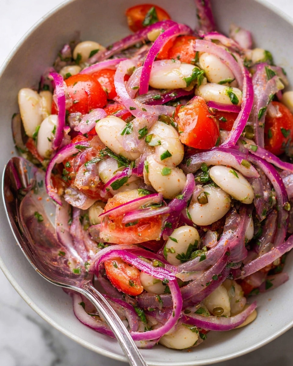 This close-up image shows a salad inside a white bowl on a white marbled surface, featuring four main layers: thin slices of bright purple-red onions twisted around the salad, creamy white large lima beans scattered evenly, small pieces of vibrant red tomatoes, and finely chopped green herbs sprinkled throughout. There is a hint of light seasoning dusted on top, giving a slightly powdery, reddish finish over the ingredients. A silver spoon with a reflective surface rests inside the bowl, nestled among the salad layers, showing a mix of beans, onion slices, and tomatoes. The ingredients look fresh and moist, with a slight shine of dressing on them photo taken with an iphone --ar 4:5 --v 7
