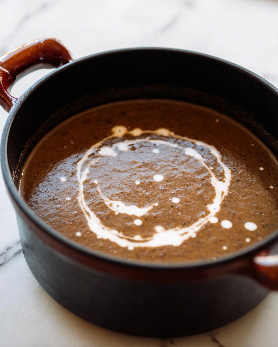 The image shows a dark brown thick soup in a black pot with a reddish-brown handle. The soup surface is smooth with visible small bits and has a swirl of white cream on top, creating a circular pattern with some drops inside it. The pot sits on a white marbled surface, and the background is softly lit, keeping the focus on the soup. Photo taken with an iphone --ar 4:5 --v 7