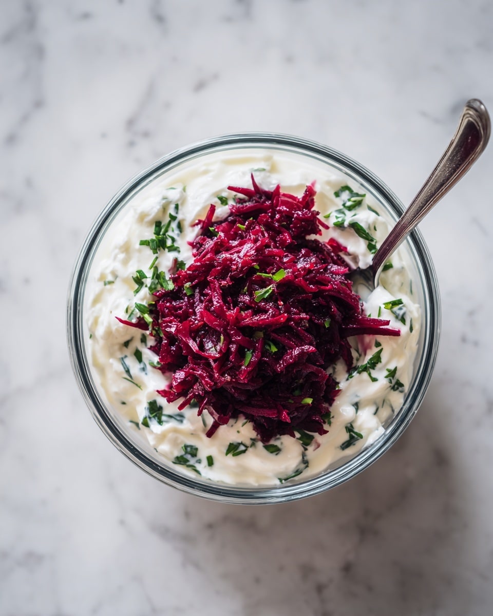 A clear glass bowl holding two layers: the bottom layer is thick and creamy white yogurt mixed with small green herb pieces, creating a textured background, and the top layer is a pile of dark red shredded beetroot placed in the center. A silver fork is partially visible on the right side, resting in the shredded beetroot. The bowl sits on a white marbled surface. photo taken with an iphone --ar 4:5 --v 7
