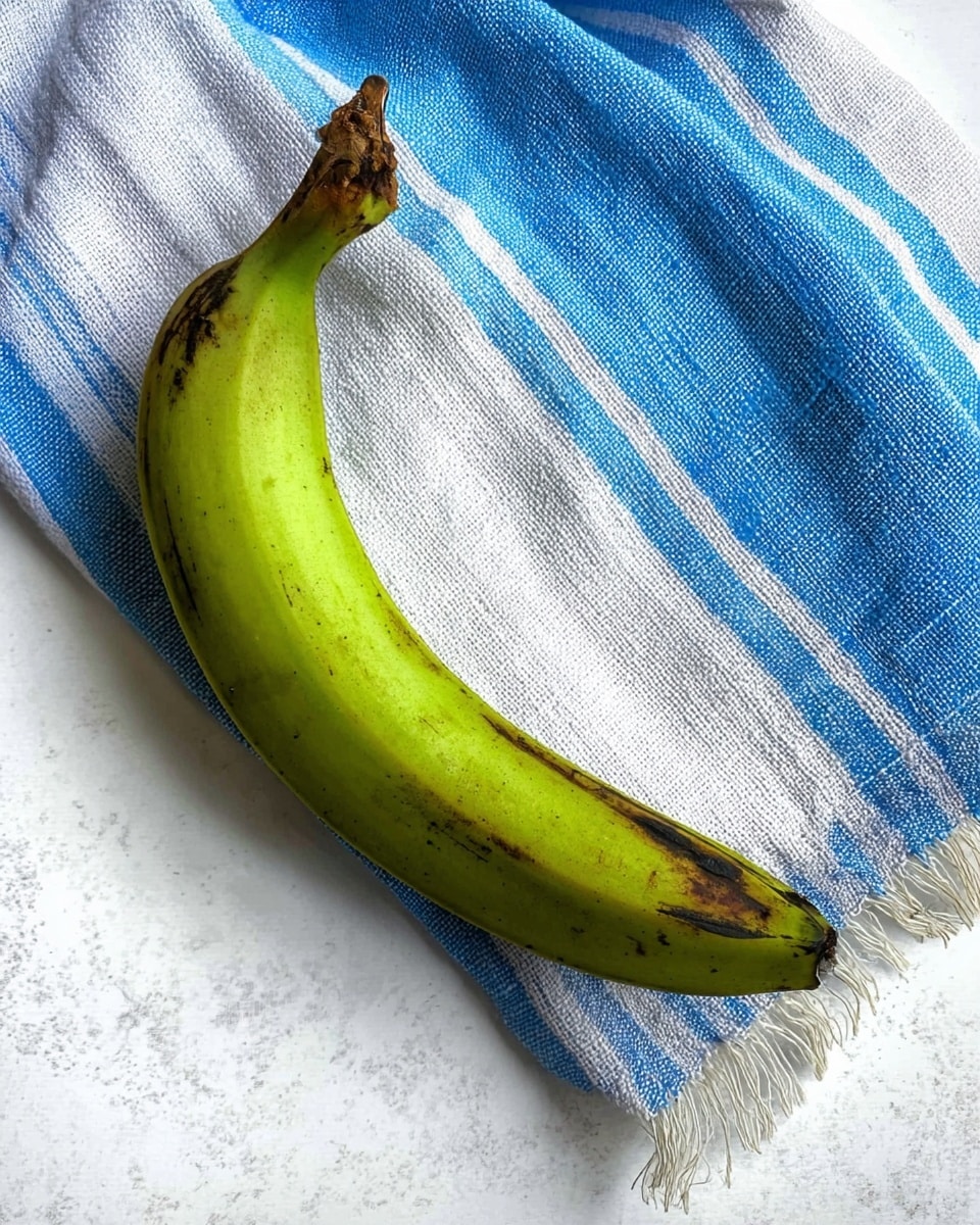 A single green plantain sits on a white marbled surface, resting partially on a blue and white striped cloth with a rough, woven texture and some frayed edges. The plantain is long and curved, with a smooth skin showing some dark spots and a darker tip at the bottom. Photo taken with an iphone --ar 4:5 --v 7