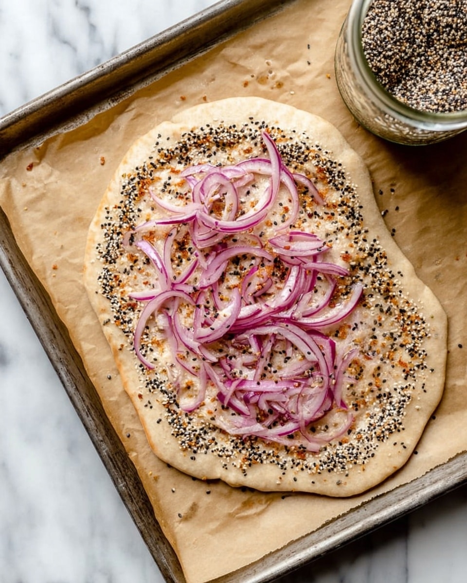 The image shows a single small flatbread on a baking tray lined with brown paper on a white marbled surface. The flatbread has one layer, a light tan dough base filled with a border of mixed black and white sesame seeds and other dry spices, creating a textured edge. On top, there is a loose pile of thinly sliced red onions arranged roughly in the center, with a slightly shiny look. The flatbread is oval-shaped and looks ready to be baked. A glass jar filled with the same seeds and spices is partially visible in the top right corner. Photo taken with an iphone --ar 4:5 --v 7