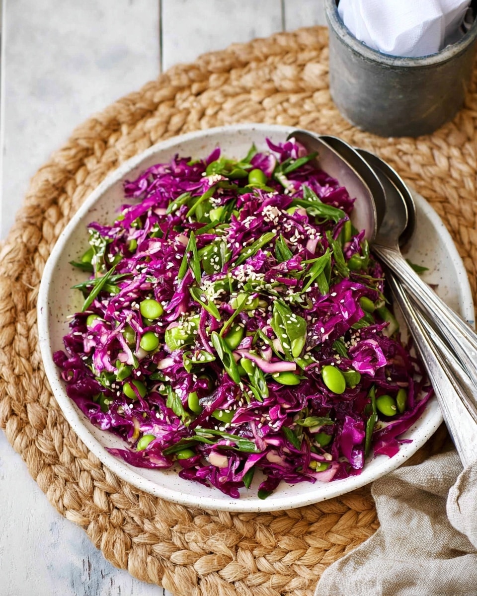 A plate filled with a colorful salad showing two main layers: deep purple shredded cabbage mixed with bright green sliced scallions and peas, sprinkled with small white sesame seeds on top. The salad is served on a white plate sitting on woven straw mat, with the plate and utensils placed on a white marbled surface. Two silver spoons rest slightly inside the edge of the plate, and a small metal container holding white napkins is visible in the background. The image shows a fresh and vibrant look with a mix of crisp textures and vivid colors. photo taken with an iphone --ar 4:5 --v 7
