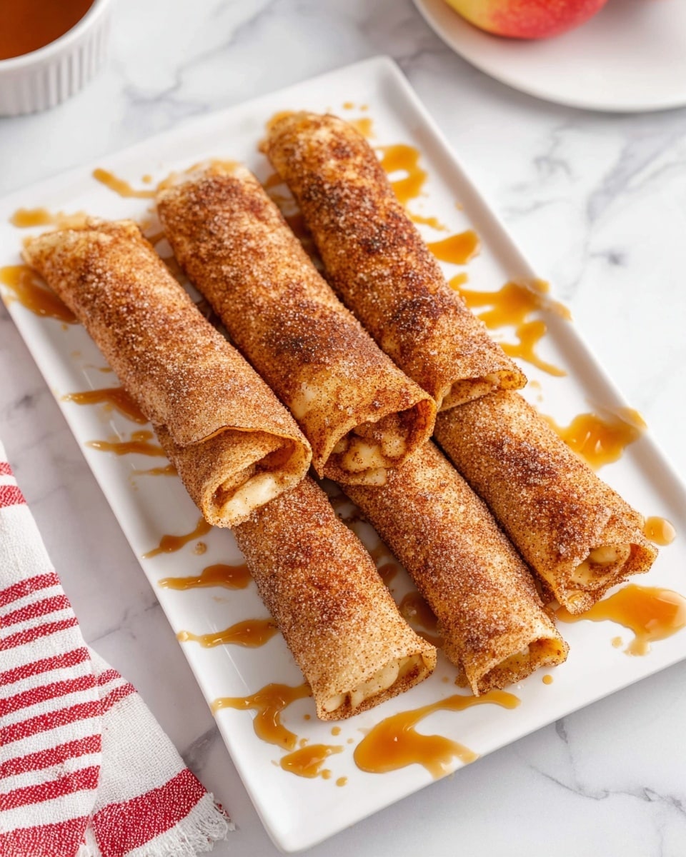 The image shows four rolled crepes placed side by side on a white rectangular plate. Each crepe is coated evenly on the outside with a cinnamon and sugar mix, giving them a brown, grainy texture. Some caramel sauce is drizzled on the plate around the crepes in a loose, artistic pattern, with a light amber color and shiny texture. The crepes appear thin and have a soft, slightly textured surface with some filling slightly visible at the ends. The plate is set on a white marbled surface with a red-striped white cloth partially visible on the left side. Photo taken with an iphone --ar 4:5 --v 7