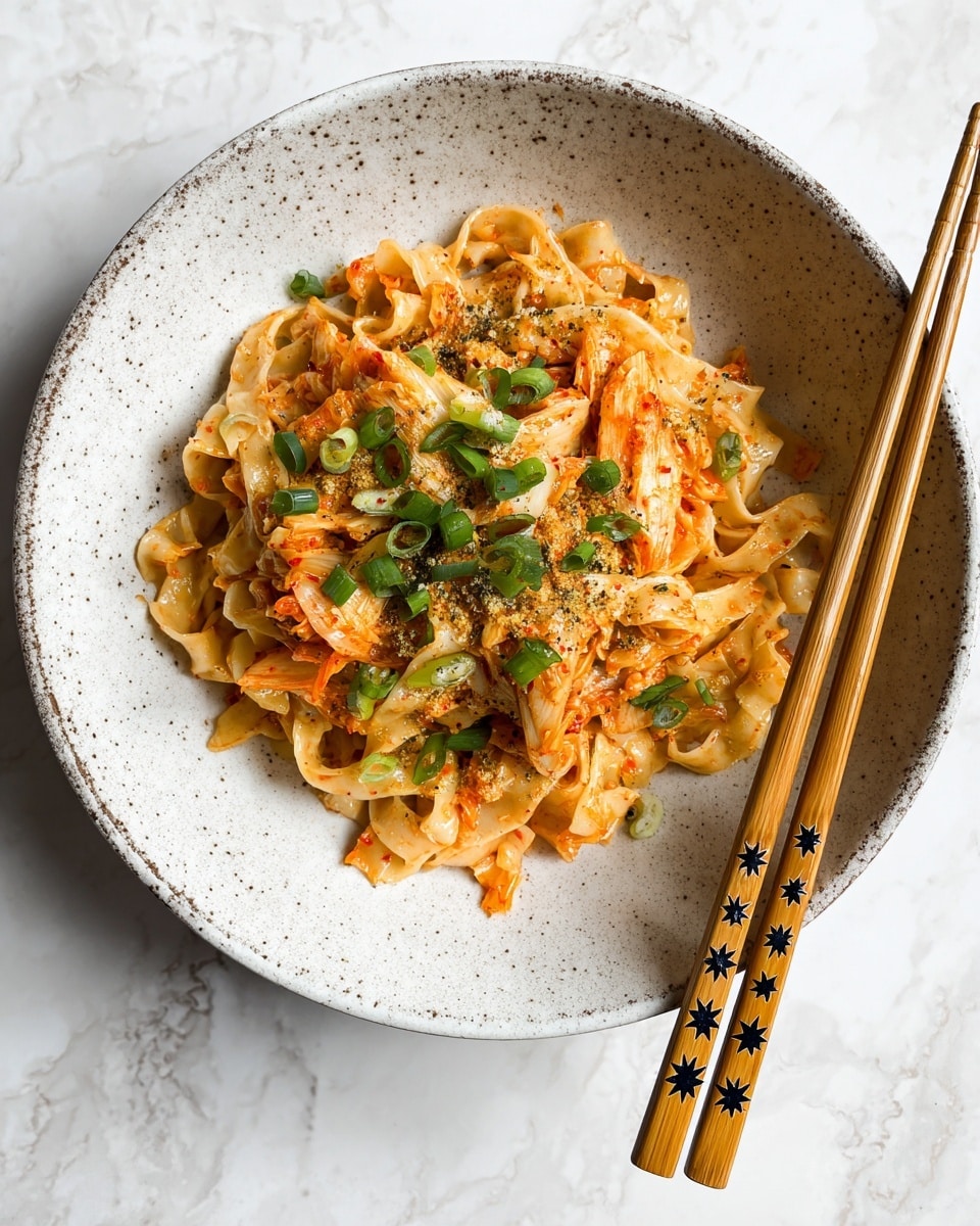 A light gray speckled white bowl holds a serving of flat, beige noodles that have a slightly rough texture, mixed with pieces of vibrant orange and white kimchi scattered on top and through the noodles. Bright green sliced scallions are sprinkled as a fresh garnish, adding contrast to the dish. The noodles are coated lightly with a powdery seasoning that gives a subtle grainy look across the surface. To the right edge of the bowl, light-colored wooden chopsticks with small decorative star patterns rest neatly. The background is a white marbled texture. photo taken with an iphone --ar 4:5 --v 7