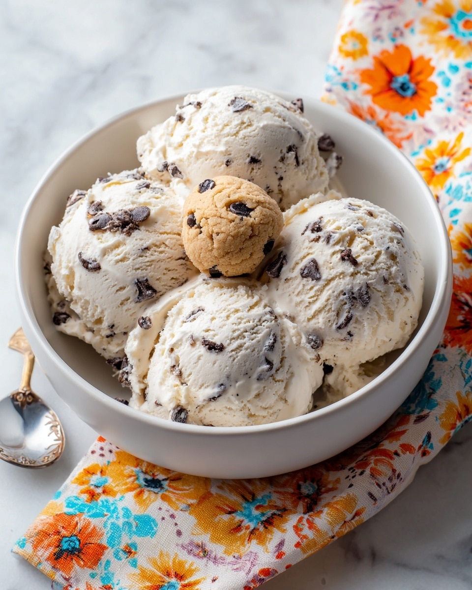A white bowl filled with several large scoops of creamy white ice cream with small dark chocolate chips mixed in, topped with two round pieces of light brown cookie dough that have visible chocolate chips, a knife with a wooden handle is stuck vertically into the cookie dough on top, the bowl is placed on a white marbled surface with a colorful, patterned cloth blurred in the background, photo taken with an iphone --ar 4:5 --v 7