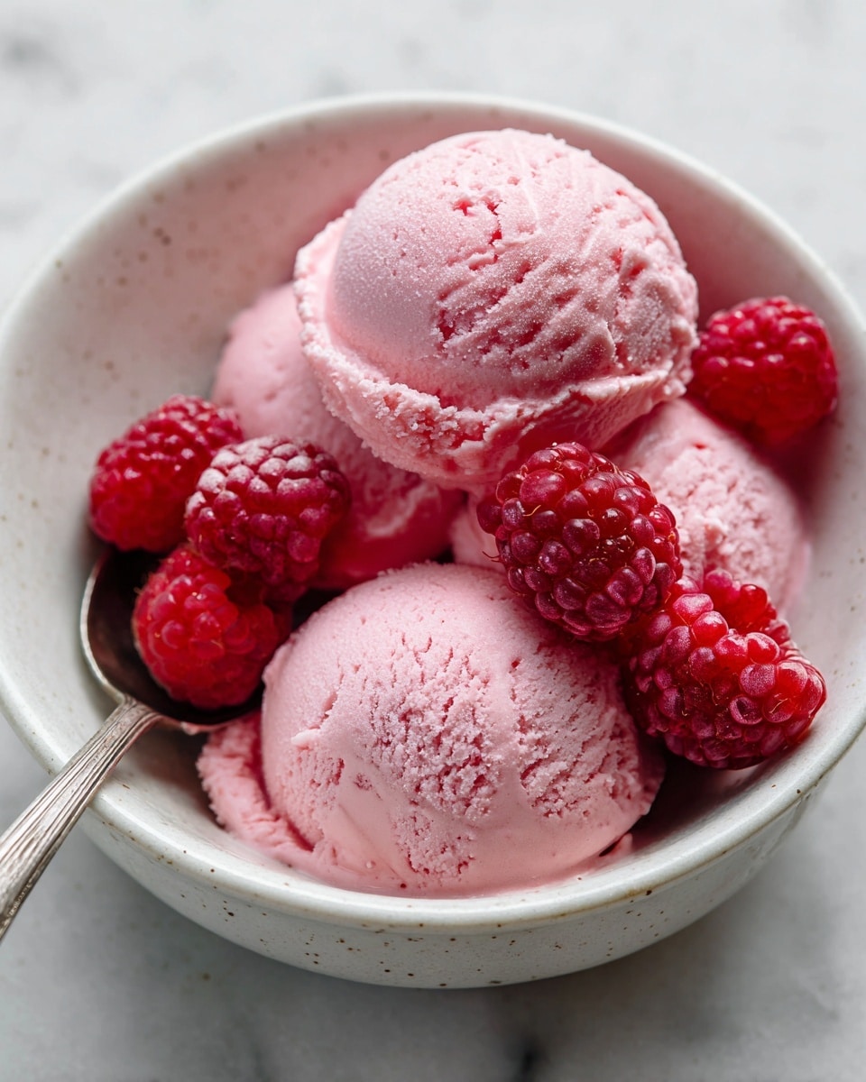 A close-up view of three scoops of pink raspberry ice cream placed in a white bowl with a light speckled interior. The ice cream has a smooth, creamy texture with slight roughness on the surface. Bright red, fresh raspberries are scattered on top and nestled between the scoops, adding vibrant color and texture contrast. A silver spoon rests inside the bowl near the edge, slightly coated with the pink ice cream. The background shows a white marbled surface. photo taken with an iphone --ar 4:5 --v 7