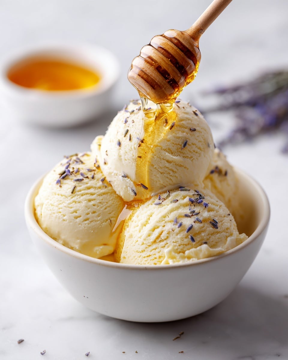 A close-up view of a white bowl placed on a white marbled surface, filled with three scoops of pale yellow creamy ice cream layered evenly. Golden honey is being drizzled over the ice cream from a wooden honey dipper held above, with the honey flowing smoothly in a thin stream. Small lavender buds are sprinkled lightly on the top scoop, adding a delicate texture and soft purple color contrast to the smooth and creamy ice cream. In the background, a small white bowl with more honey is slightly blurred, enhancing the focus on the main bowl of ice cream. Photo taken with an iphone --ar 4:5 --v 7