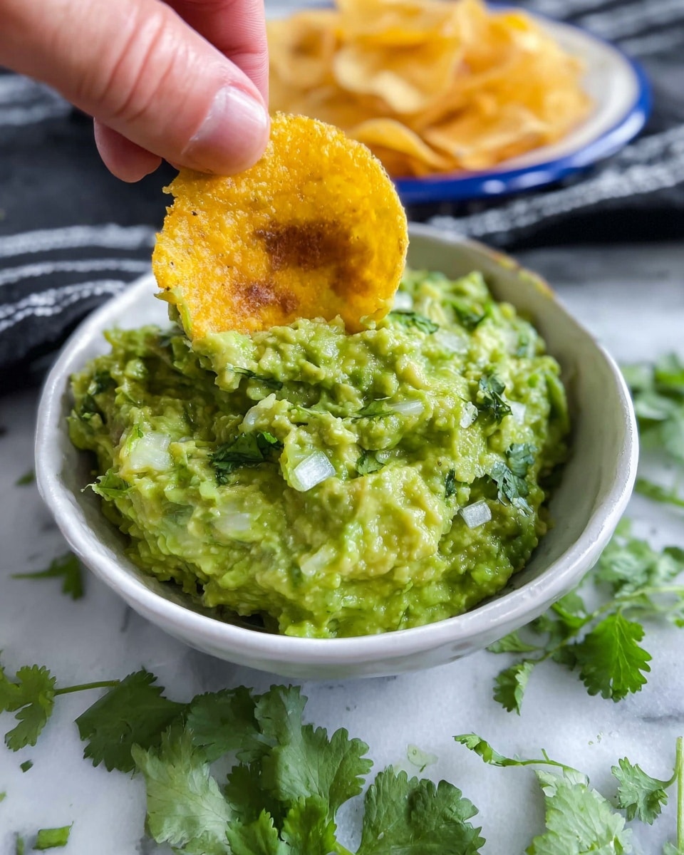 The image shows a close-up of a white bowl filled with chunky green guacamole that has visible bits of onion and cilantro mixed in. On top of the guacamole, a golden-yellow crispy round chip with a textured, ruffled edge is being dipped by a woman's hand. Around the bowl, fresh cilantro leaves are scattered on a white marbled surface. In the background, there is a white plate with a blue rim holding more of the same chips, placed on a folded black and white striped cloth. Photo taken with an iphone --ar 4:5 --v 7