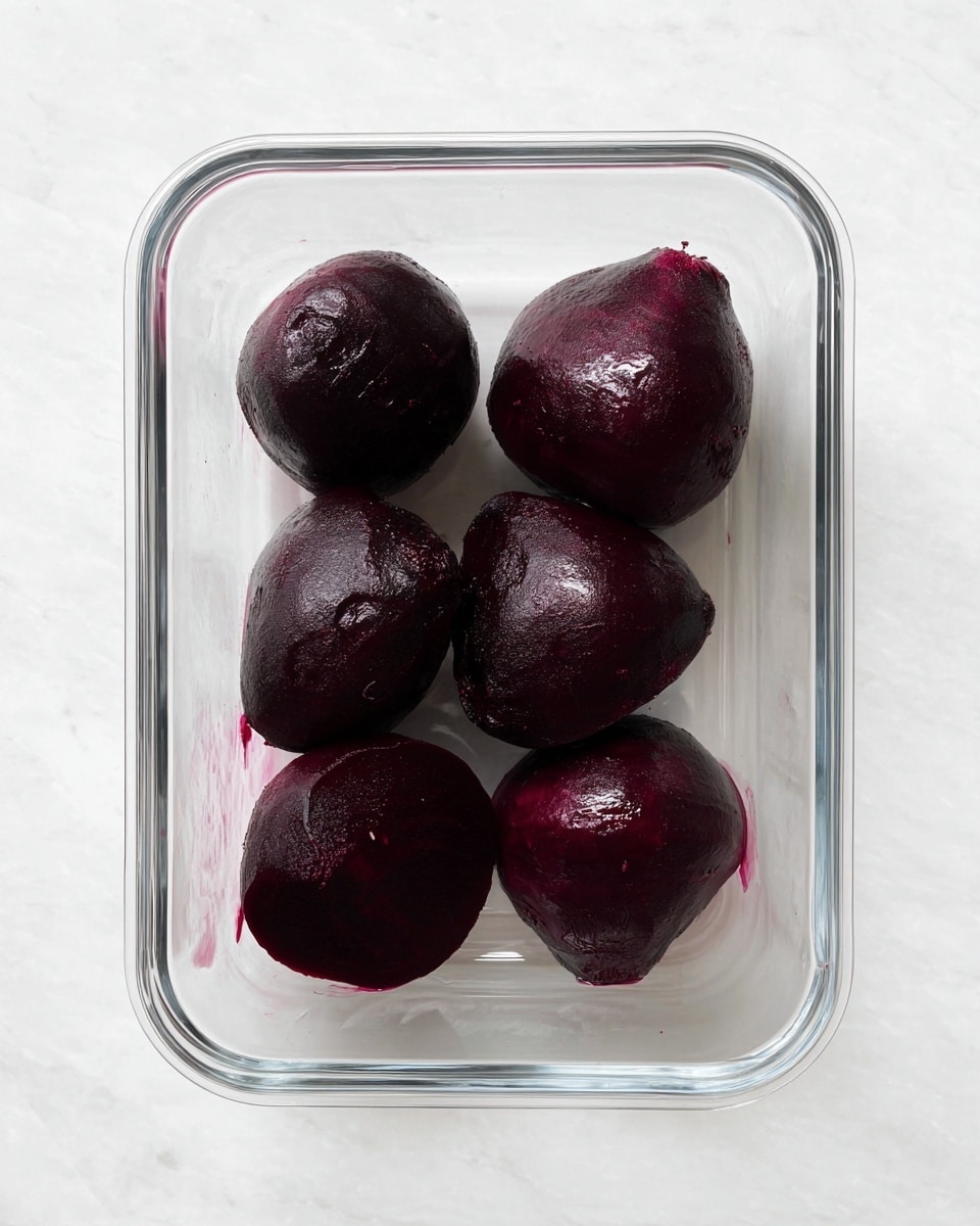The image shows six peeled dark purple beets placed in a clear rectangular glass container. The beets have a smooth, slightly shiny texture, varied in size and shape, some with rounded tops and others with more irregular surfaces. The container rests on a white marbled surface, giving a clean and simple background look. No other items are in the picture, focusing solely on the beets and the container. Photo taken with an iphone --ar 4:5 --v 7