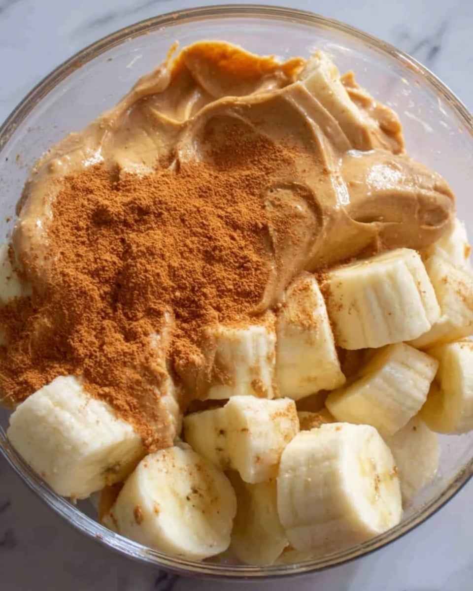 The image shows a close-up view of a glass bowl filled with three layers of ingredients. At the bottom, there are large pieces of peeled banana, creamy and pale yellow in color, roughly cut and filling most of the bowl. On top of the bananas, there is a thick layer of light brown peanut butter with a smooth, slightly textured surface. Finishing the bowl, a fine, reddish-brown powder, likely cinnamon, is sprinkled on one side, creating a contrast in color and texture against the peanut butter and banana. The bowl is placed on a white marbled surface. photo taken with an iphone --ar 4:5 --v 7
