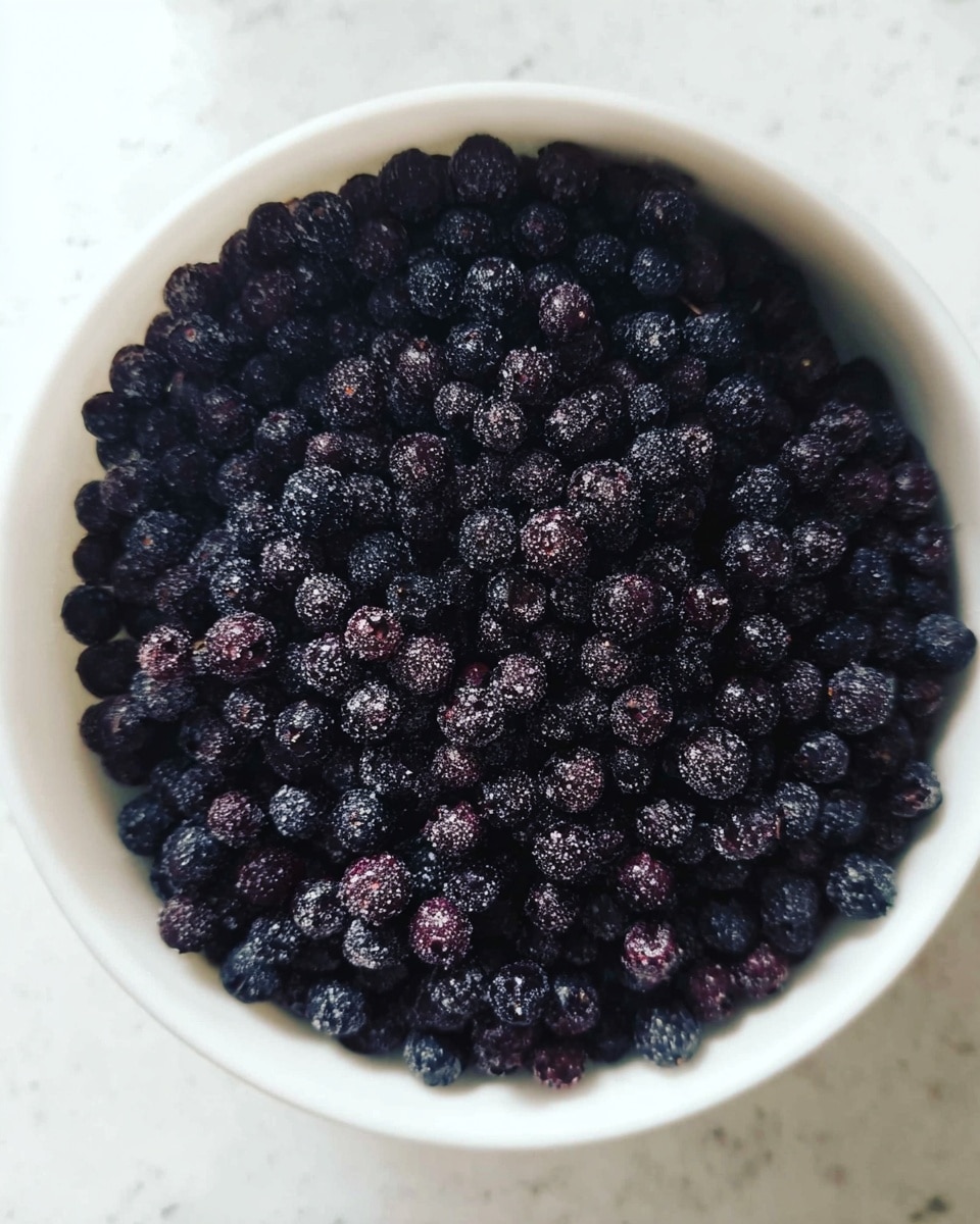 A close-up image shows a white bowl filled with a large amount of small dark purple berries that have a matte and slightly frosty texture, tightly packed to fill the bowl. The berries vary slightly in size, creating a natural, uneven surface. The bowl rests on a white marbled surface. photo taken with an iphone --ar 4:5 --v 7