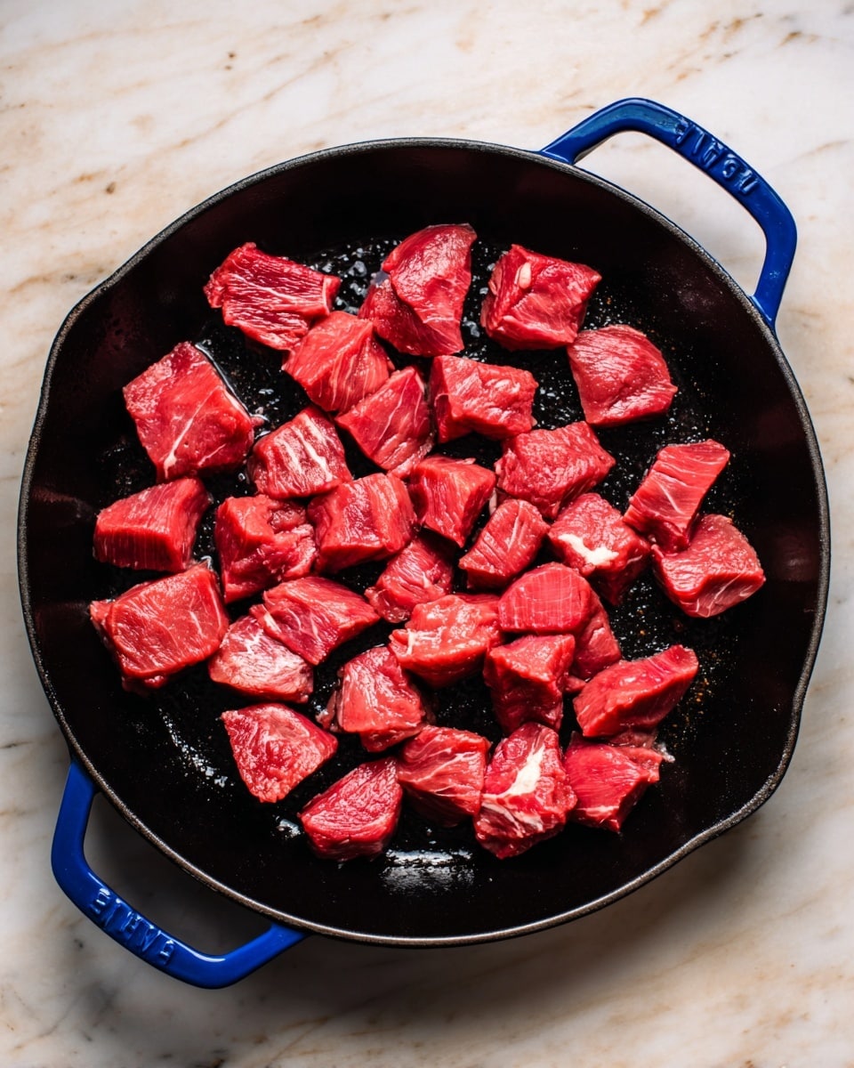 The image shows a black cast iron pan with a blue handle on a white marbled surface. Inside the pan, there are many pieces of raw red meat, cut into medium-sized cubes and spread evenly across the pan. Some pieces have small streaks of white fat. The pan itself has a slightly shiny, oily texture where the meat touches it, and the background is a clean white marble texture. photo taken with an iphone --ar 4:5 --v 7