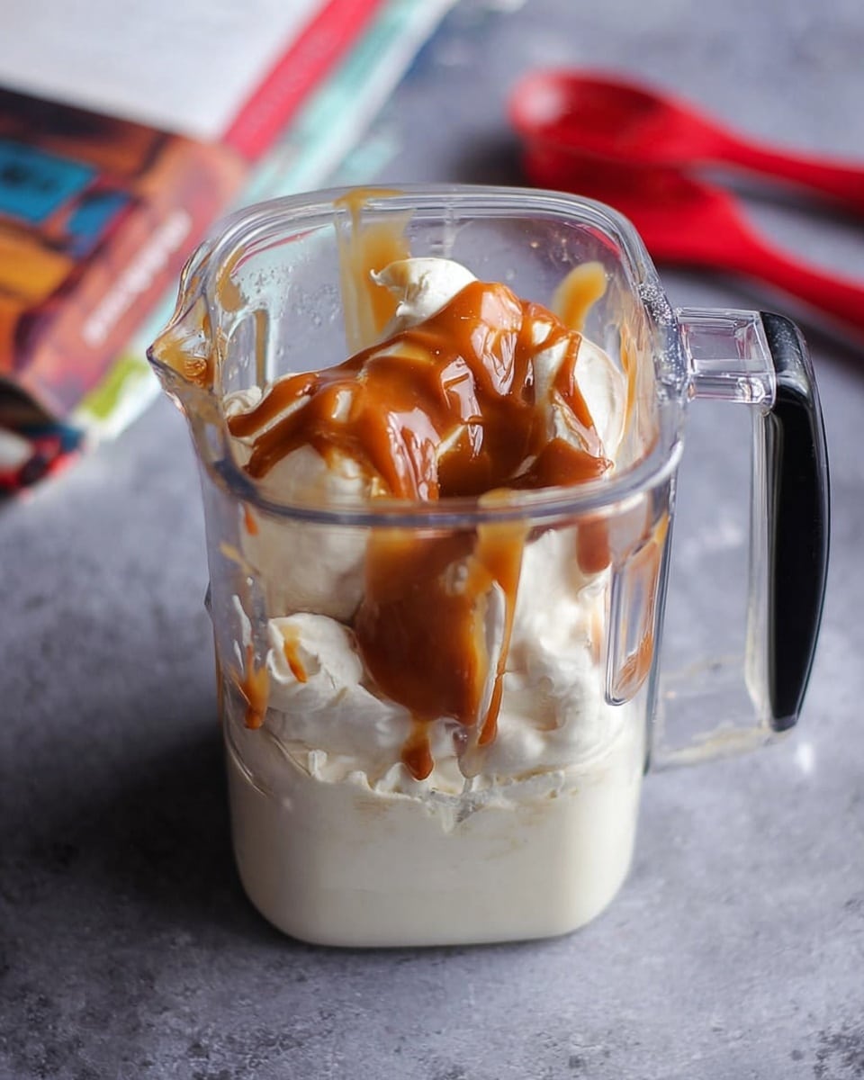 A clear blender jar shows layers of thick white cream as the base with smooth, rich brown caramel sauce drizzled unevenly over it. The jar has a black handle on the right side and sits against a soft gray surface with a red-handled object and a colorful open book in the background. The light catches the shiny caramel sauce, giving it a glossy texture, while the cream looks fluffy and dense. photo taken with an iphone --ar 4:5 --v 7