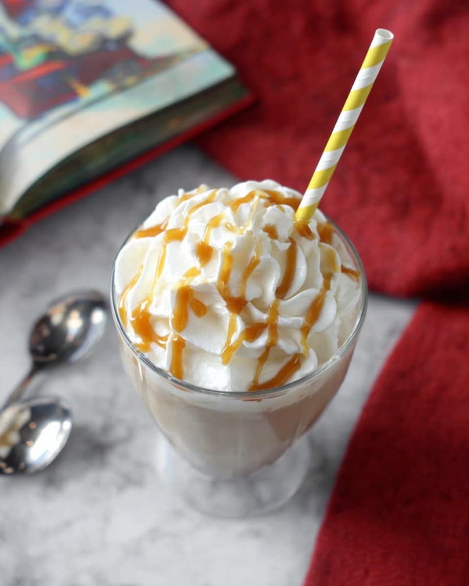 A clear glass filled with a light brown creamy drink is topped with a thick layer of white whipped cream that has been swirled to create soft peaks. On top of the whipped cream is a drizzle of caramel sauce in loose lines, adding warm amber color and shine. A yellow and white striped paper straw is placed in the glass, standing straight up from the center of the whipped cream. The glass is set on a white marbled surface with a red cloth partially visible to the side, and the blurred edge of an open colorful book is near the top left of the image. Two shiny silver spoons lie nearby on the white marbled background. photo taken with an iphone --ar 4:5 --v 7