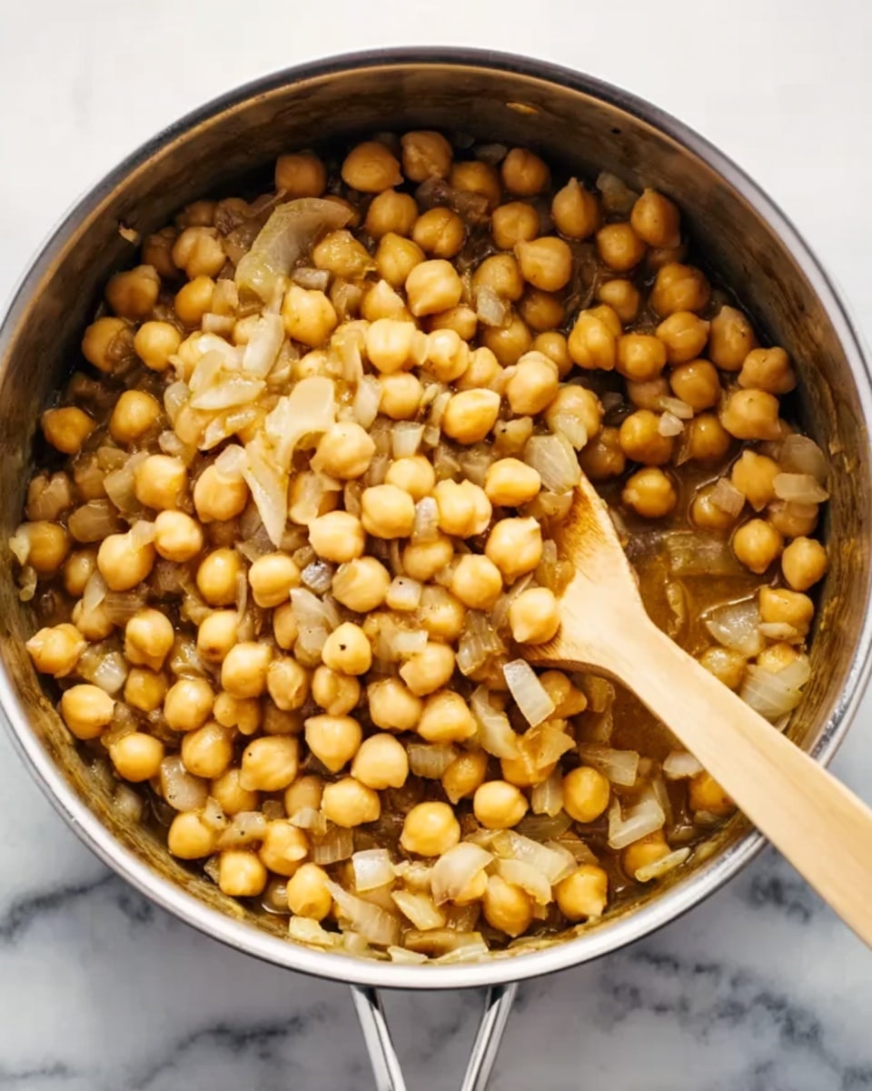 The image shows a close-up of a silver pot filled with cooked chickpeas mixed with small pieces of onion and light brown sauce. The chickpeas are round and creamy yellow with a soft, smooth texture, layered evenly throughout the pot. The onion pieces are translucent white, scattered lightly among the chickpeas. A wooden spoon is placed inside the pot, partially covered with the chickpea mixture. The pot sits on a white marbled surface. Photo taken with an iphone --ar 4:5 --v 7