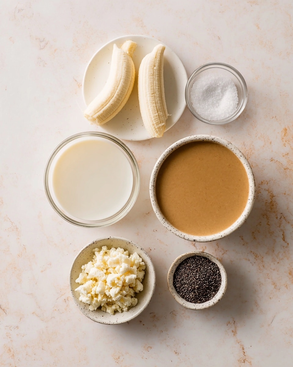 The image shows six small bowls and dishes on a white marbled surface, each with different ingredients. At the bottom left is a clear bowl filled with a white liquid. To the right, a small white dish holds two peeled bananas. Above that is a round white plate with a pile of small, white, crumbly pieces. Above the plate, there is a white bowl filled with a brown creamy sauce. To the right of the bowl, a white speckled dish holds black chia seeds. At the top center, a small clear glass bowl contains white coarse salt. The arrangement is simple and clean, with soft lighting highlighting the textures. Photo taken with an iphone --ar 4:5 --v 7