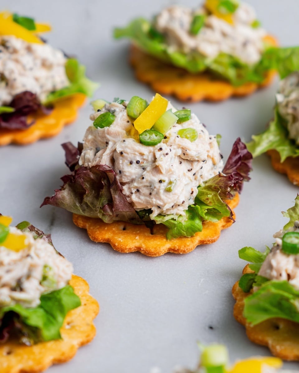 The image shows small cracker snacks arranged on a white marbled surface, each with three visible layers. The base layer is an orange, crispy, round cracker with scalloped edges. On top of the cracker is a layer of mixed green and purple lettuce leaves, giving a fresh, leafy texture. The top layer is a generous dollop of creamy white tuna salad mixed with black pepper, small pieces of celery, and bits of yellow pepper, garnished with slices of green onion and small yellow pepper pieces on top. The focus is on one cracker in the center, with others slightly blurred in the background. photo taken with an iphone --ar 4:5 --v 7