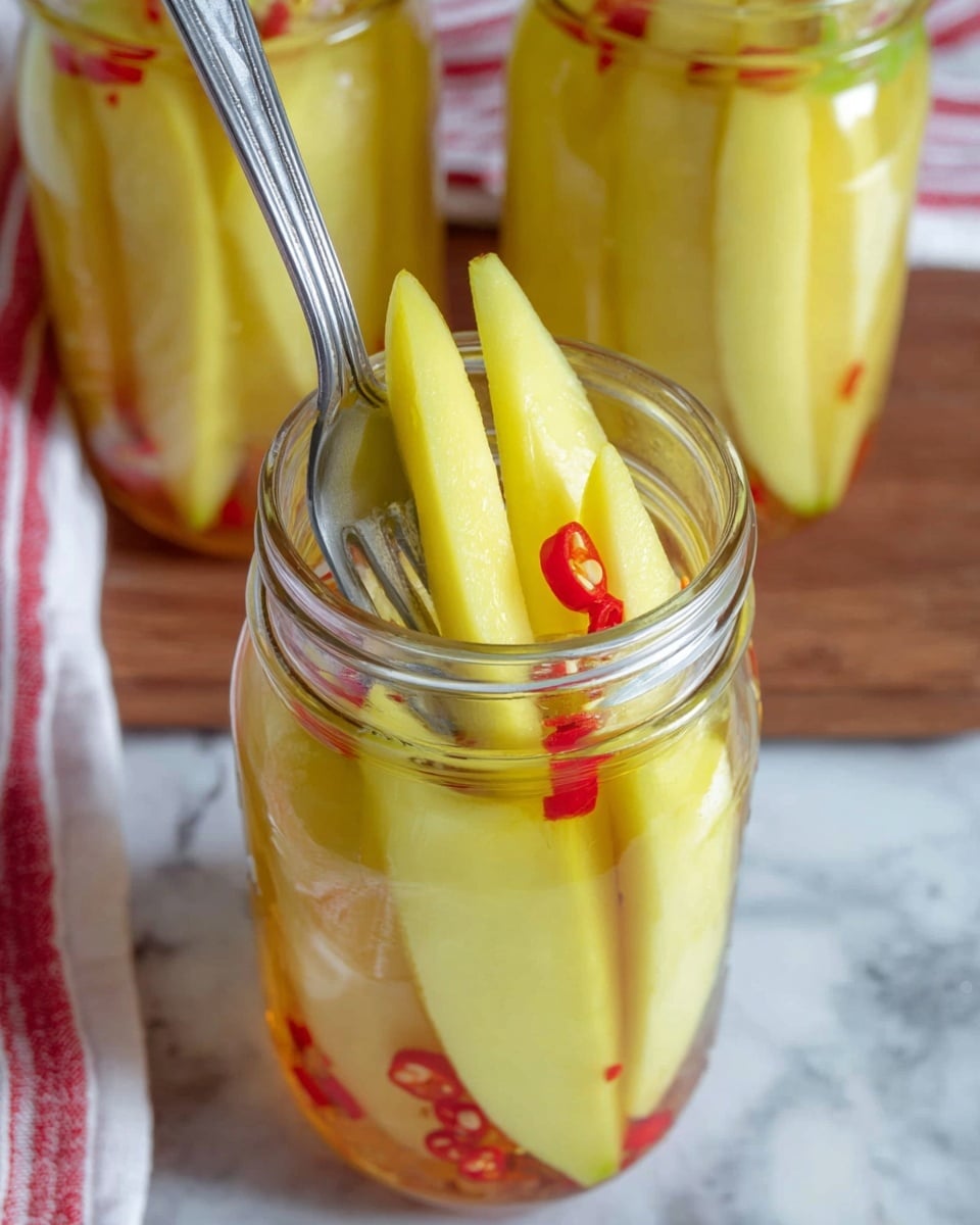 A clear glass jar filled with tall, pale yellow slices of mango, standing upright and surrounded by small red chili pieces and some seeds in a clear liquid. A silver metal fork is shown gently lifting one mango slice out of the jar. In the background, there are two more similar jars with the same mango and chili slices inside, placed on a wooden surface with a white marbled texture underneath. Part of a red and white striped cloth is visible on the left side. photo taken with an iphone --ar 4:5 --v 7