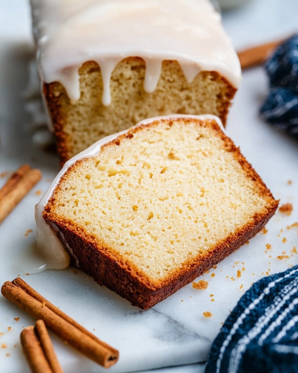A thick slice of light golden brown cake sits on a white marbled surface, topped with a thin, shiny layer of white glaze that drips down the sides. The cake has a soft, fluffy texture with tiny air holes throughout. The crust is a darker brown, forming a firm frame around the lighter inner cake. Close by, two cinnamon sticks lie flat, adding a warm brown color contrast. Part of a larger loaf, coated with the same white glaze, is visible on the right side. A striped dark blue cloth is slightly visible in the background. Photo taken with an iphone --ar 4:5 --v 7