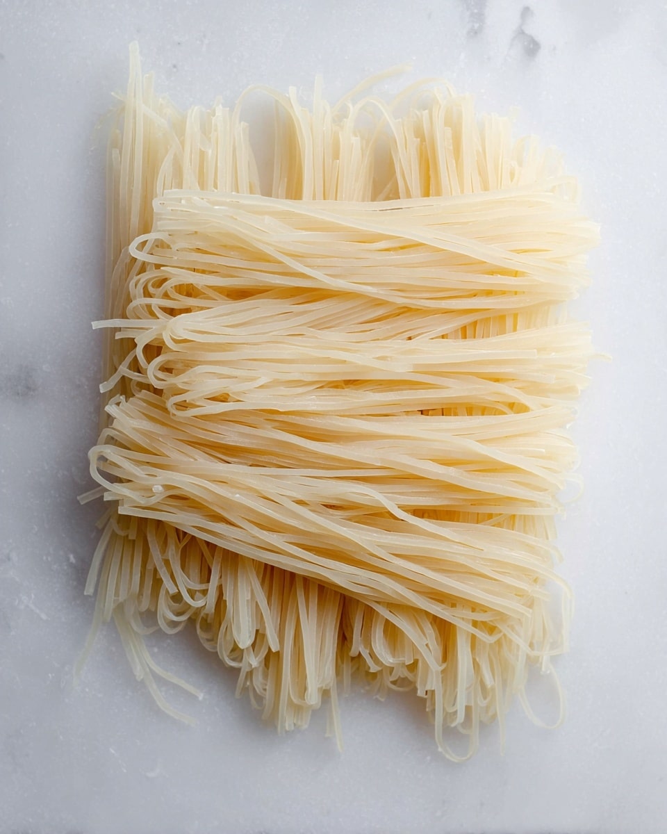 A pile of raw, thin rice noodles spread out in a loose rectangular shape on a white marbled surface. The noodles are pale yellow and semi-transparent, with a smooth and slightly shiny texture. They are arranged side by side, with some strands overlapping and others standing slightly upright or bending, showing their flexibility and softness. The noodles vary slightly in length but are mostly uniform, creating a delicate layered effect. Photo taken with an iphone --ar 4:5 --v 7