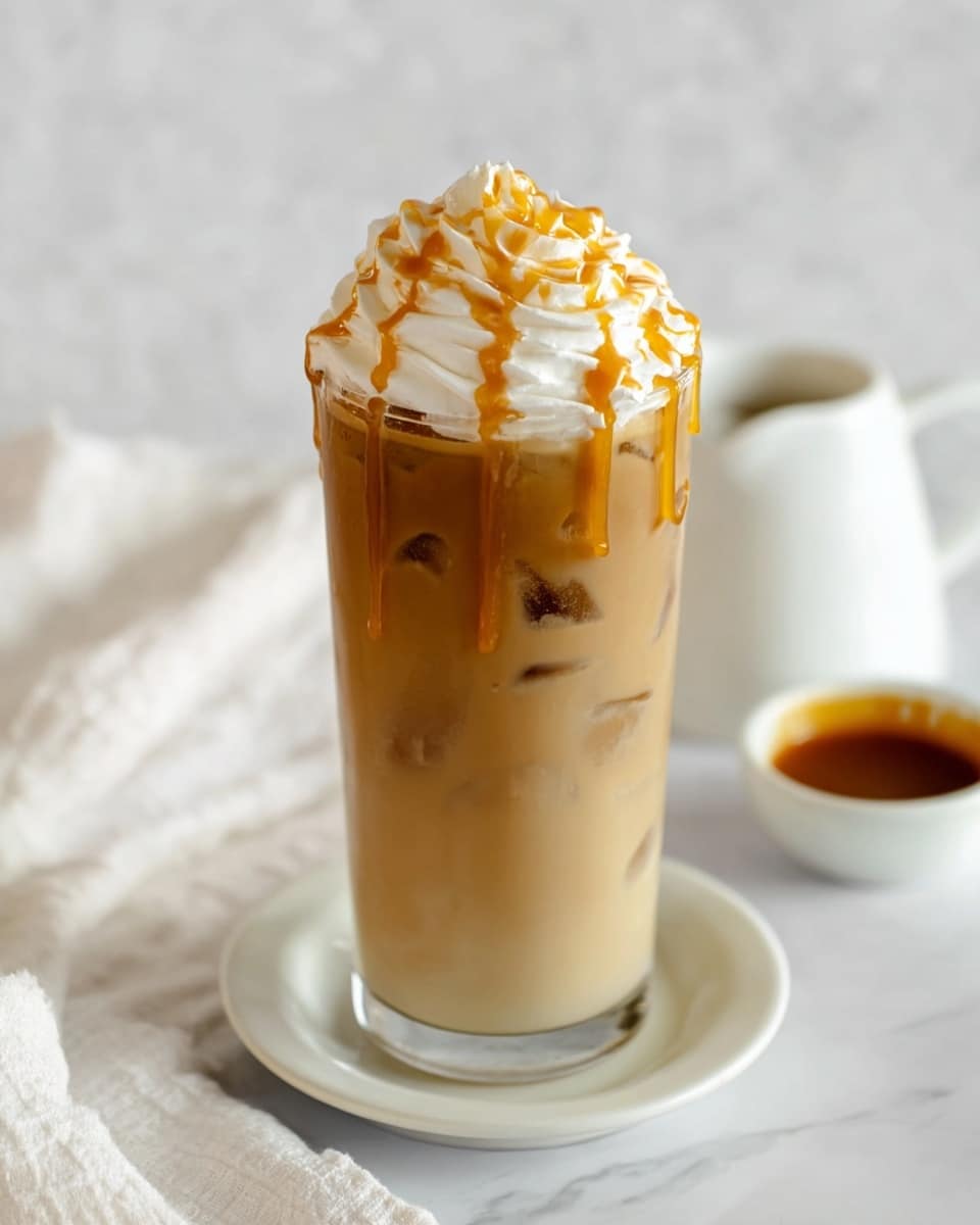 A tall clear glass holds a creamy light brown iced coffee filled with ice cubes visible inside. On top, there is a thick swirl of white whipped cream, drizzled with golden caramel sauce that gently drips down the sides of the cream. The glass sits on a white saucer against a white marbled surface, with a small white bowl of caramel sauce and a white cloth nearby, creating a soft and inviting look. Photo taken with an iphone --ar 4:5 --v 7