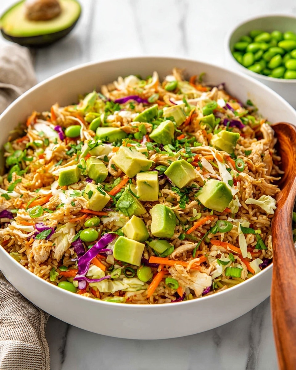 A large white bowl filled with a colorful mixed rice dish layered with light brown cooked rice, shredded white and purple cabbage, orange carrot strips, bright green edamame beans, and cubed avocado pieces on top. There are also some shredded light brown chicken pieces spread evenly throughout the dish, along with small green herbs and thinly sliced green onions sprinkled across. A wooden spoon rests inside the bowl on the right side. The bowl is placed on a white marbled surface with a small white bowl of green edamame beans blurred in the background. Photo taken with an iphone --ar 4:5 --v 7