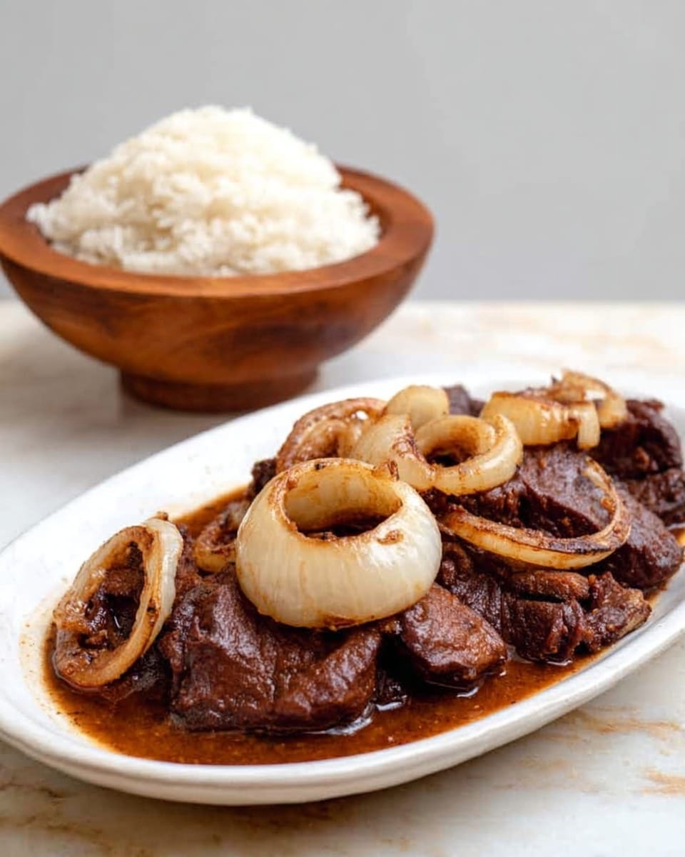 A white oval plate holds about five pieces of dark brown cooked meat topped with soft cooked white onion rings mixed with the meat juices. Behind the plate, there is a small wooden bowl filled with white rice. The whole scene is set on a white marbled surface with a plain light background. Photo taken with an iphone --ar 4:5 --v 7