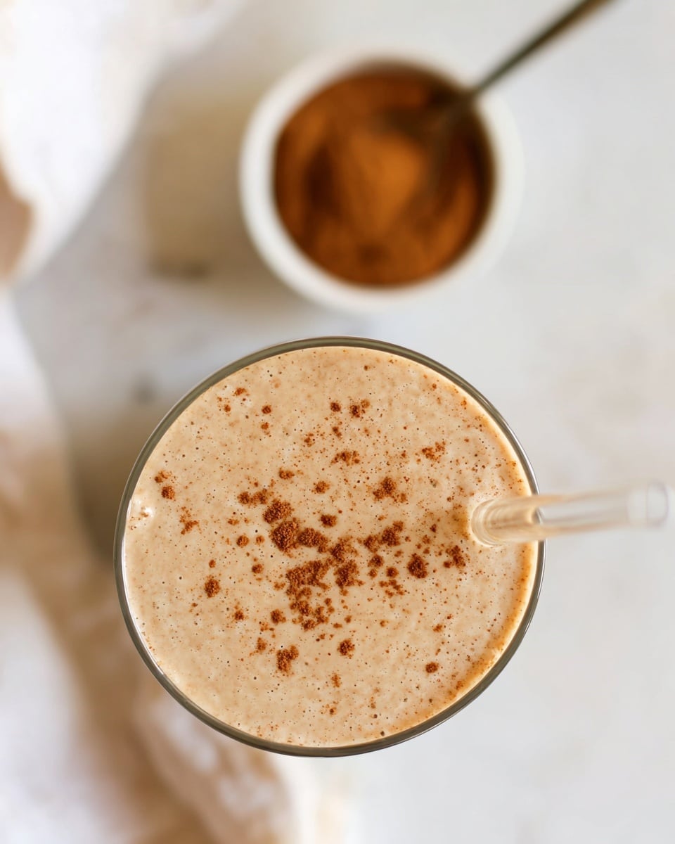 A top view of a creamy light beige smoothie in a clear tall glass, with small cinnamon or spice specks sprinkled on its smooth surface. A transparent straw is placed inside the glass on the right side. In the background, a blurred white bowl filled with brown cinnamon powder and a spoon rests on a white marbled surface. The overall look is soft and inviting with warm tones. photo taken with an iphone --ar 4:5 --v 7