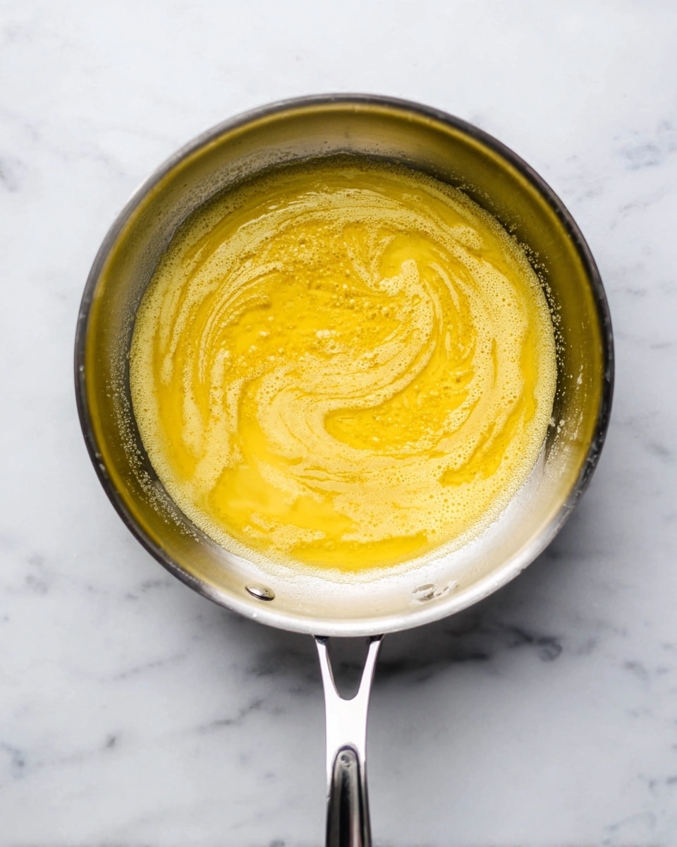 A shiny silver pan with a thick handle in the center holds melted golden yellow butter with light foam and a slightly grainy texture on top, swirling gently inside. The pan sits on a white marbled surface that gives a clean and bright look, and the smooth yellow butter contrasts with the shiny metal edges of the pan. Photo taken with an iphone --ar 4:5 --v 7